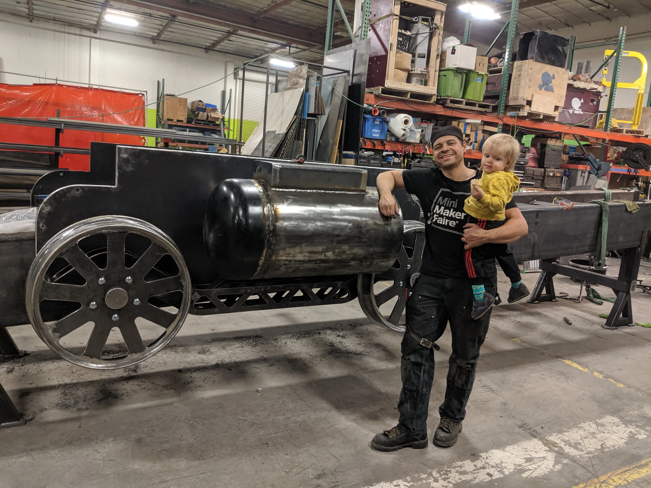 Andy Tibbetts holding a child beside Lawn Dart, showing the scale of the locomotive sculpture